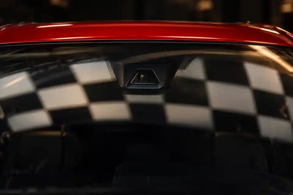 Illustration — boîtier de la caméra d’aide à la conduite derrière le pare-brise d’une voiture rouge, un panneau de calibrage à damier s’y reflète.