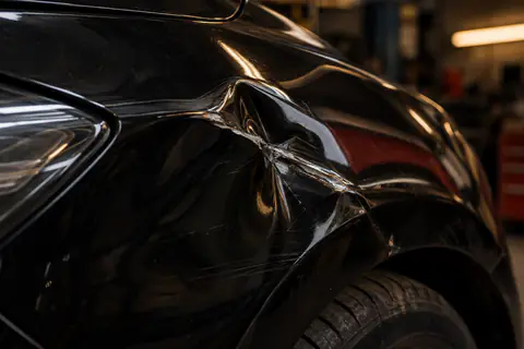 Illustration — aile avant de voiture enfoncée en gros plan, tôle noire marquée par un choc, reflet rouge.